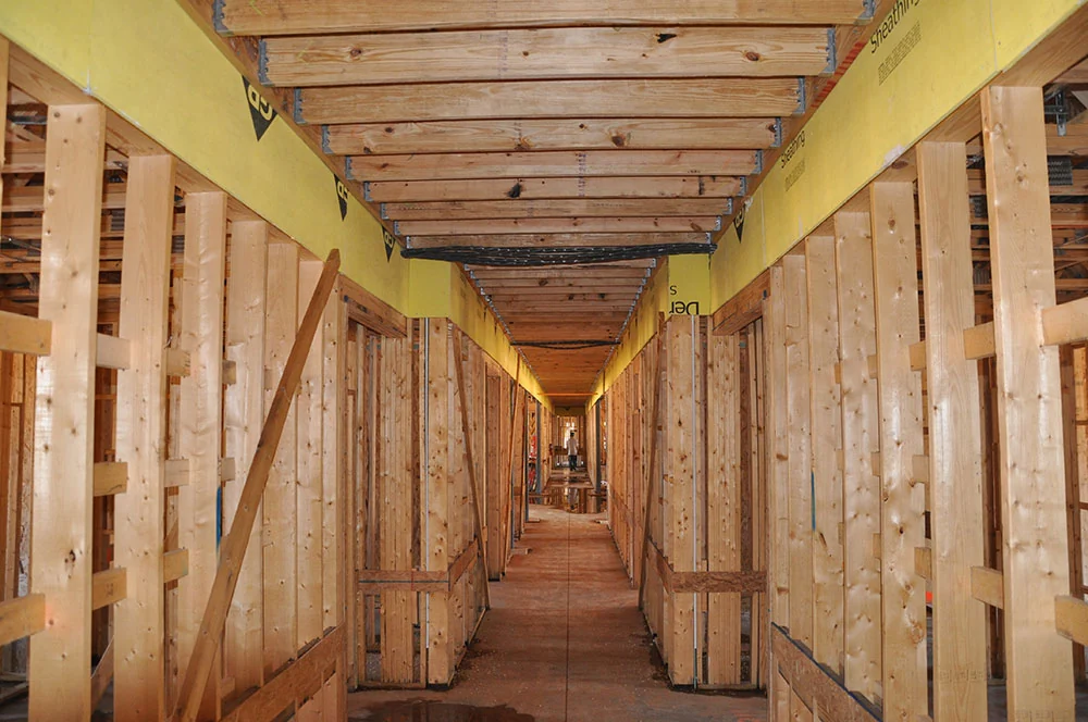 Wood-framed interior hallway with exposed studs, a partial ceiling grid, and an opening to the exterior.